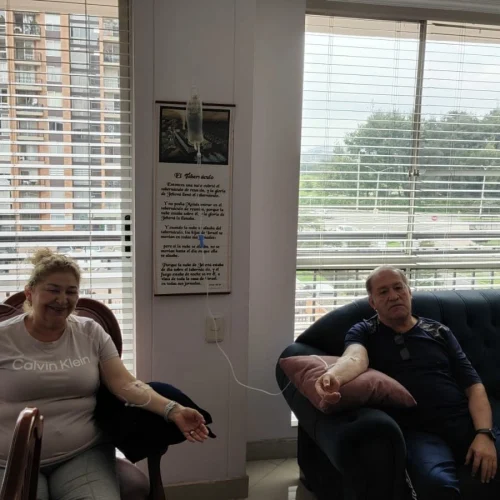 Dos personas sentadas en sillones junto a una ventana, recibiendo sueroterapia a través de vías intravenosas conectadas a un frasco de suero colgado en la pared.