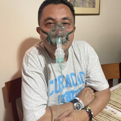 Hombre sentado en una mesa con los brazos cruzados, usando una mascarilla de oxígeno conectada a un tubo de nebulización, en un entorno doméstico.
