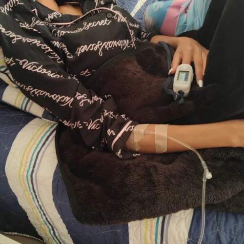 Mujer recostada en una cama con pijama negra estampada, recibiendo sueroterapia a través de una vía intravenosa en el brazo y con un oxímetro colocado en un dedo, en un entorno doméstico.