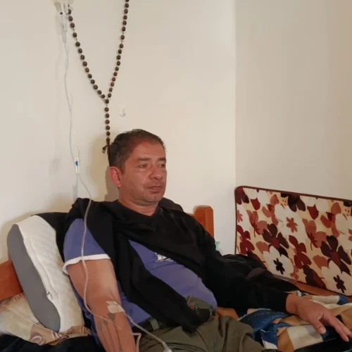 Juan Carlos Melo sentado en una cama recibiendo sueroterapia intravenosa en el brazo izquierdo, con bolsa de suero colgada en la pared y un rosario alrededor.