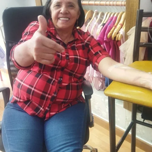 Mujer sonriente con camisa de cuadros rojos y negros sentada en una silla, recibiendo sueroterapia en el brazo derecho, levantando el pulgar en señal de aprobación.